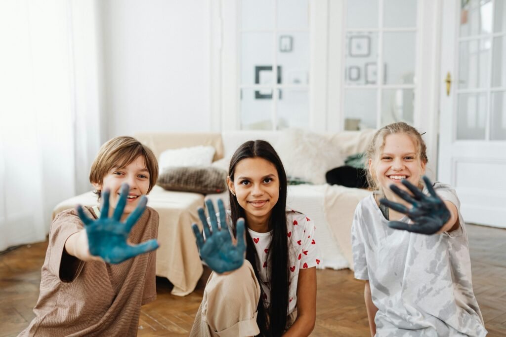 Three smiling teenagers showing hands covered in blue paint at home.
